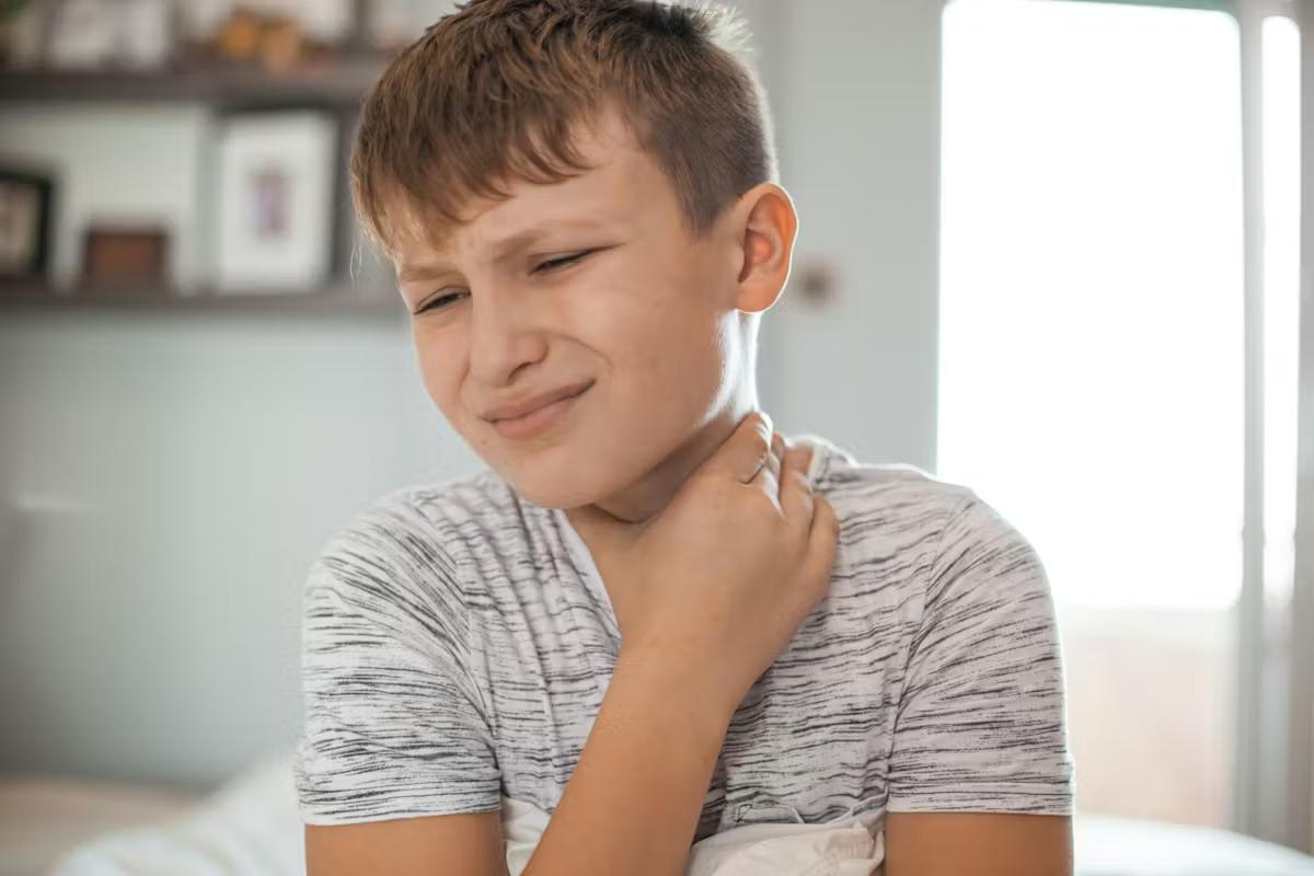 School-aged boy sitting at home with a sore throat and holding his neck.